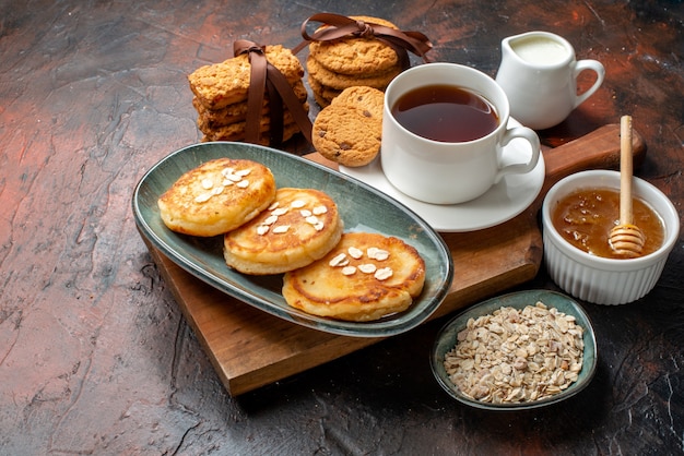 Vista frontal de panqueques frescos una taza de té negro sobre una tabla de cortar de madera galletas de miel apiladas leche sobre una superficie oscura