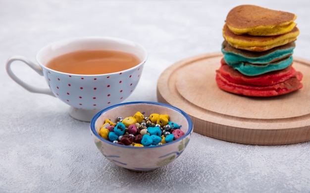 Vista frontal de panqueques multicolores sobre un soporte con chocolates de colores y una taza de té sobre un fondo blanco.