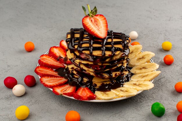 Vista frontal de panqueques de postre de frutas con fresas rojas en rodajas de chocolate y plátanos dentro de un plato blanco sobre el gris