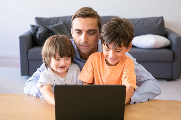 Vista frontal de papá abrazando a sus hijos y viendo películas en la computadora portátil. Dos niños pequeños encantadores sentados a la mesa con el padre, mirando la pantalla y sonriendo. Concepto de paternidad, infancia y tecnología digital.