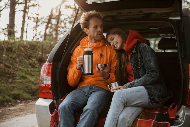 Vista frontal de la pareja disfrutando de una bebida caliente en el maletero del coche