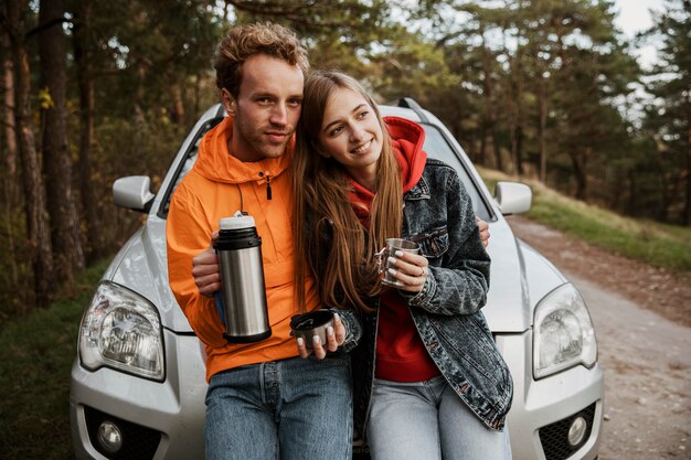 Vista frontal de la pareja disfrutando de una bebida caliente mientras está sentado en el capó del coche al aire libre