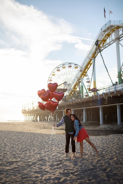 Vista frontal de una pareja joven abrazada en la playa mientras sostiene globos en forma de corazón