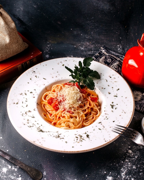 Vista frontal pasta cocida junto con hojas verdes frescas y tomates rojos dentro de un plato blanco sobre el escritorio gris