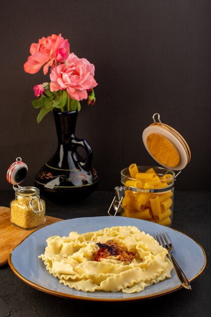 Una vista frontal de la pasta de masa cocida sabrosa salada dentro de la placa azul redonda con flores dentro de la jarra en la alfombra diseñada y el escritorio oscuro cocina italiana