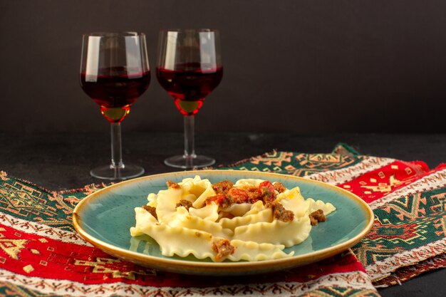 Una vista frontal de la pasta de masa cocida sabrosa salada dentro de la placa verde redonda con copas de vino en la alfombra diseñada y escritorio oscuro