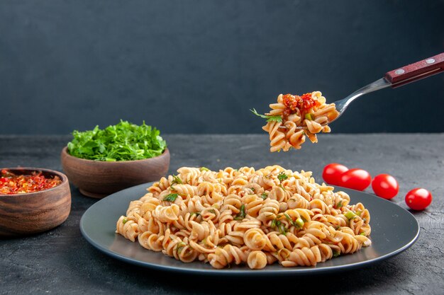 Vista frontal de la pasta rotini en un plato y en un tenedor verduras picadas en un tazón de tomates cherry sobre una superficie oscura aislada