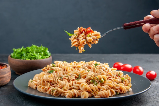 Vista frontal de la pasta rotini en el tenedor de la placa en mano femenina verduras picadas en un tazón de tomates cherry sobre superficie gris