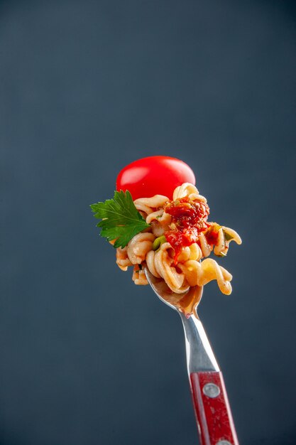 Vista frontal de pasta rotini con tomate cherry en horquilla sobre superficie oscura aislada con espacio de copia