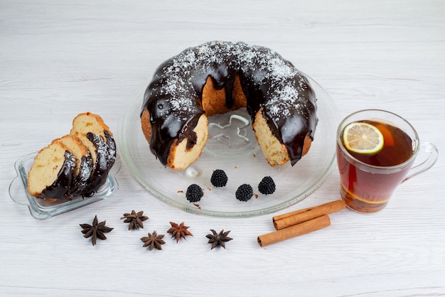 Una vista frontal de pastel de chocolate entero y en rodajas con canela y té sobre el fondo blanco pastel de galleta
