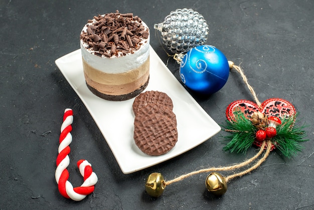 Vista frontal de pastel de chocolate y galletas en placa rectangular blanca juguetes de árbol de Navidad en la oscuridad