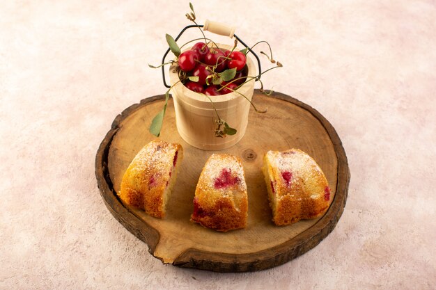 Una vista frontal pastel de frutas horneado delicioso en rodajas con cerezas rojas en el interior y azúcar en polvo en un escritorio de madera con cerezas frescas en rosa