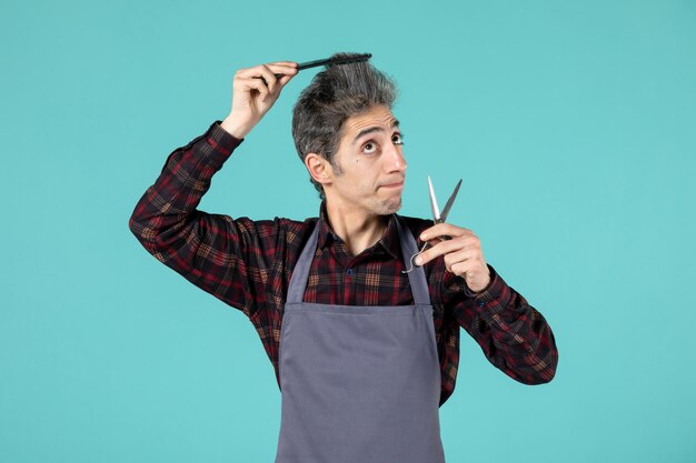 Vista frontal del peluquero masculino concentrado vistiendo delantal gris y sosteniendo una tijera peinando su cabello sobre fondo de color azul suave