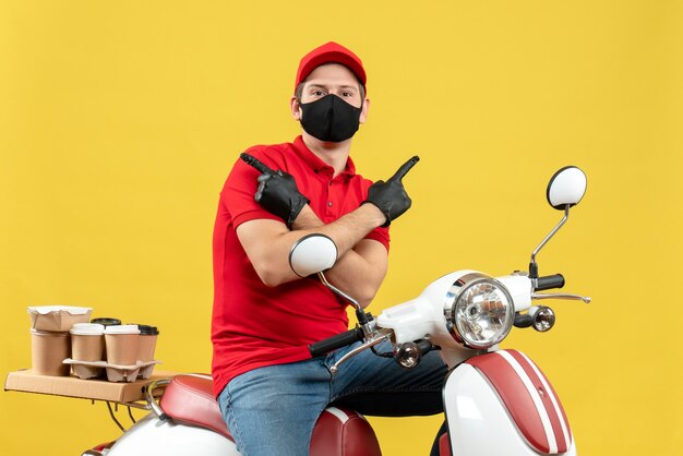 Vista frontal del pensamiento adulto joven vistiendo blusa roja y guantes de sombrero en máscara médica entregando orden sentado en scooter apuntando a ambos lados sobre fondo amarillo