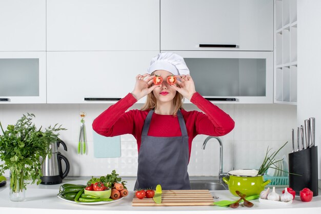 Vista frontal pequeña cocinera en delantal poniendo tomates delante de sus ojos