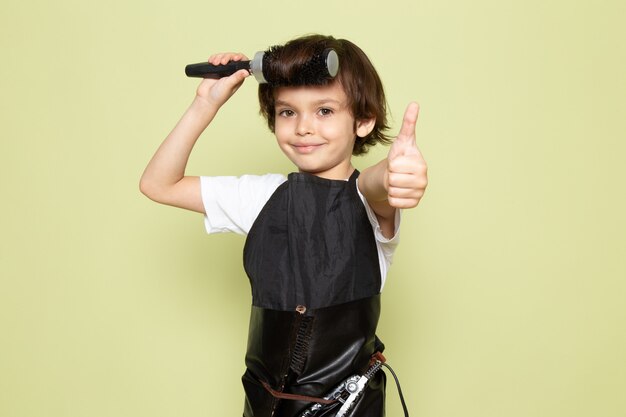 Una vista frontal pequeña peluquería adorable niño en capa negra posando