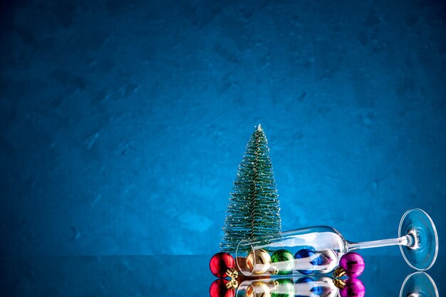 Vista frontal pequeñas bolas de Navidad esparcidas desde la copa de vino mini árbol de Navidad en la superficie azul oscuro