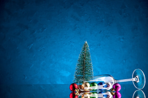 Vista frontal pequeñas bolas de Navidad esparcidas desde la copa de vino mini árbol de Navidad en la superficie azul oscuro