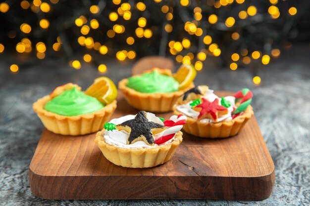 Vista frontal pequeñas tartas en la tabla de cortar en luces de Navidad de superficie oscura aislada