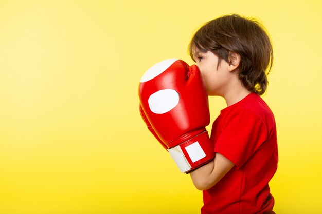 Una vista frontal pequeño niño en camiseta roja y guantes rojos en la pared amarilla