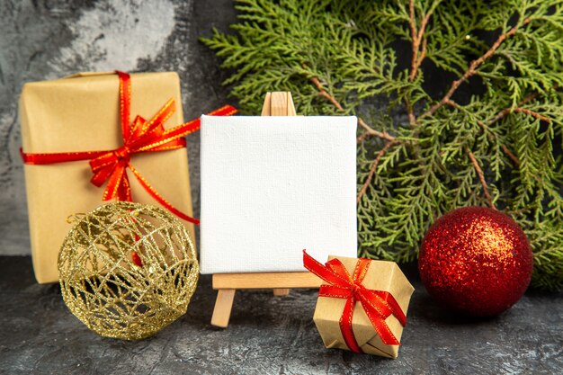 Vista frontal pequeño regalo atado con cinta roja mini lienzo en caballete de madera rama de pino bolas de navidad en gris