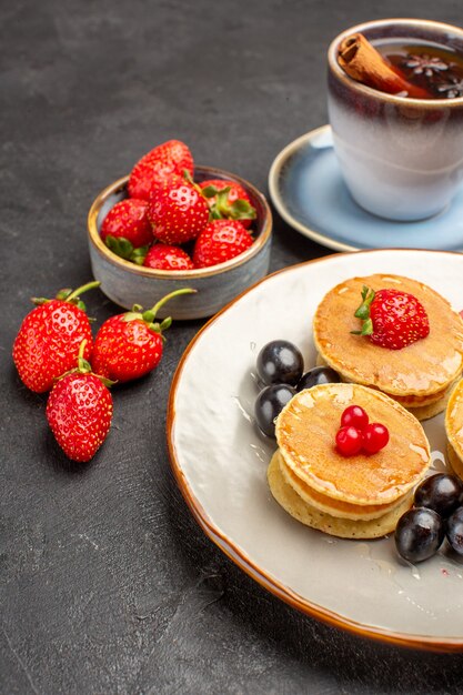 Vista frontal pequeños y deliciosos panqueques con frutas y una taza de té en frutas de pastel de superficie gris