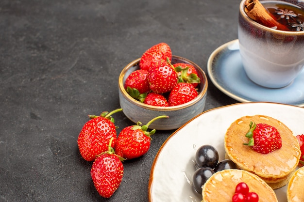 Vista frontal pequeños panqueques deliciosos con frutas y una taza de té en pastel de superficie gris oscuro