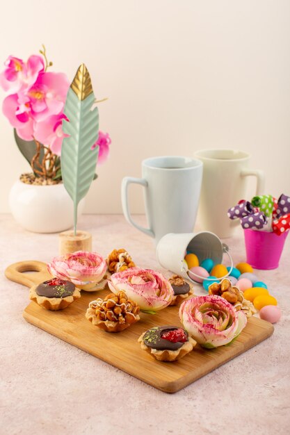 Una vista frontal de pequeños pasteles de chocolate con flores y plantas en el escritorio rosa