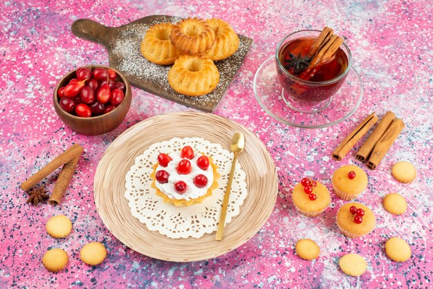 Vista frontal de pequeños pasteles con crema fresca y frutas frescas junto con canela y té en la galleta de superficie brillante