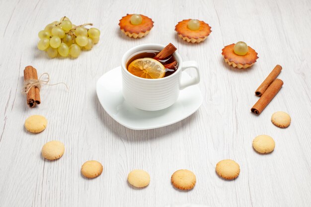 Vista frontal pequeños pasteles deliciosos con uvas taza de té y galletas en el escritorio blanco pastel de frutas galletas postre dulce té