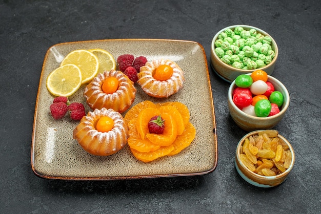 Vista frontal de pequeños pasteles con rodajas de limón, mandarinas y dulces en el espacio oscuro