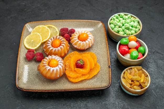 Vista frontal de pequeños pasteles con rodajas de limón, mandarinas y dulces en el espacio oscuro