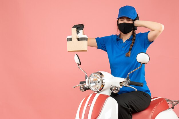 Vista frontal de la persona de entrega femenina agotada con máscara médica y guantes sentado en scooter con órdenes sobre fondo melocotón pastel