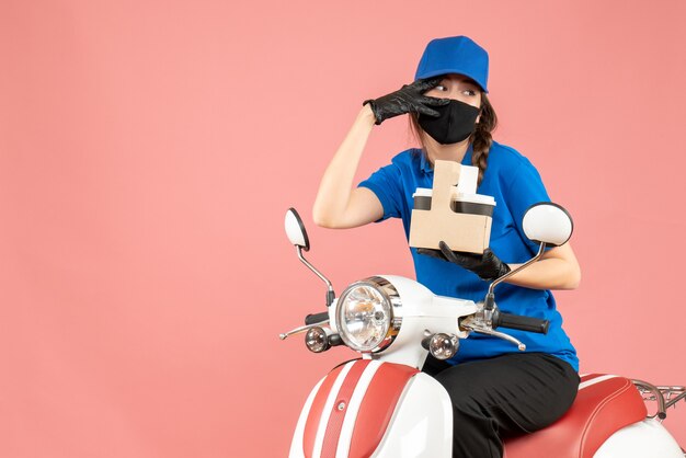 Vista frontal de la persona de entrega femenina en cuestión con máscara médica y guantes sentado en scooter entregando pedidos sobre fondo de melocotón pastel