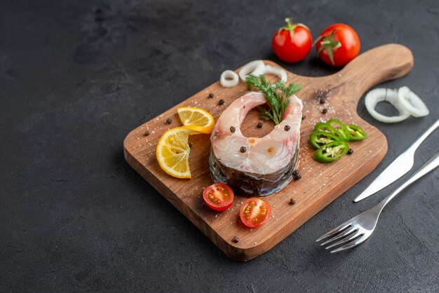 Vista frontal de pescado crudo y verduras frescas picadas rodajas de limón especias sobre una tabla de madera en el lado izquierdo sobre una superficie angustiada negra