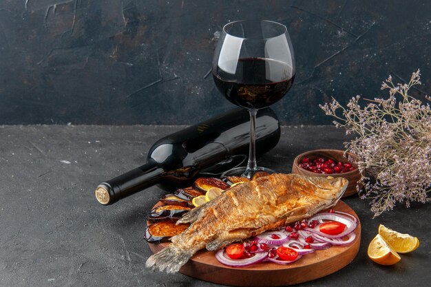 Vista frontal de pescado frito berenjenas fritas cebolla cortada sobre tabla de servir de madera botella de vino y vidrio sobre fondo oscuro