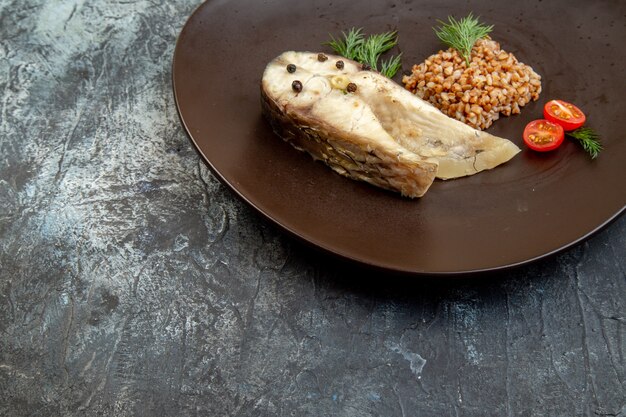 Vista frontal de pescado hervido y harina de trigo sarraceno servido con tomates verdes en un plato negro sobre una superficie de hielo con espacio libre