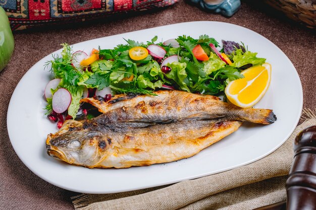 Vista frontal de pescado a la parrilla con una ensalada de verduras y hierbas con una rodaja de limón