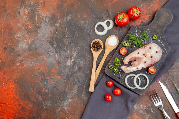 Vista frontal de los pescados crudos y los tomates verdes de cebolla pimiento en una tabla de cortar negra sobre cubiertos de toalla en una superficie de color mixto