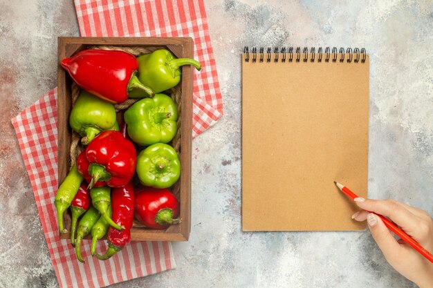 Vista frontal pimientos rojos y verdes pimientos picantes en caja de madera mantel a cuadros blanco rojo cuaderno lápiz rojo en mano de mujer sobre fondo desnudo