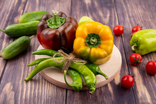 Vista frontal de pimientos en tabla de cortar con pepinos y tomates en superficie de madera