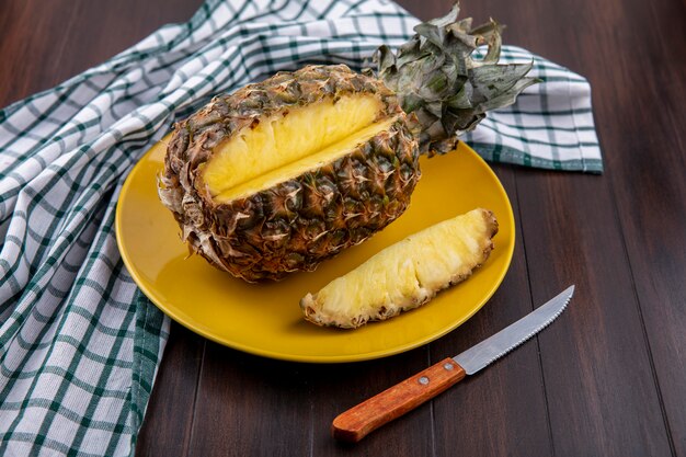Vista frontal de la piña con una pieza cortada de fruta entera en un plato sobre tela escocesa con un cuchillo sobre una superficie de madera