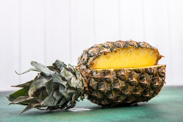 Vista frontal de piña con una pieza cortada de fruta entera en superficie verde y superficie blanca