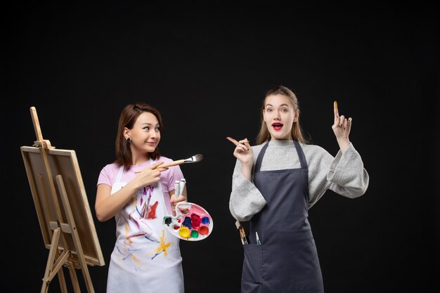 Vista frontal de la pintora dibujando en el caballete con otra mujer en la pared negra foto color arte imagen artista pintar