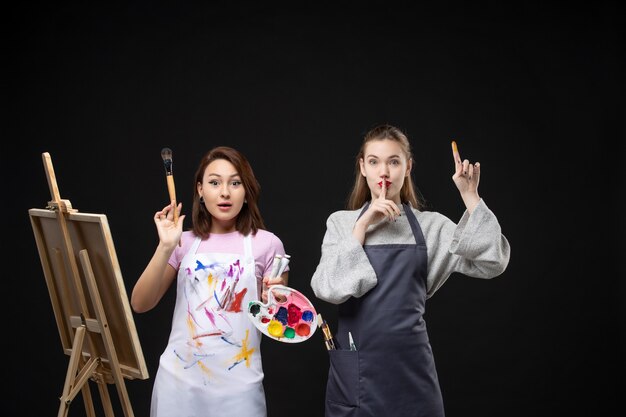Vista frontal pintora dibujando en caballete con otra mujer en la pared negra foto color arte imagen artista trabajo de pintura