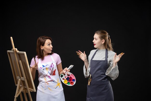 Vista frontal pintora dibujando en caballete con otra mujer en pared negra foto color arte imágenes artista pintura trabajo dibujar