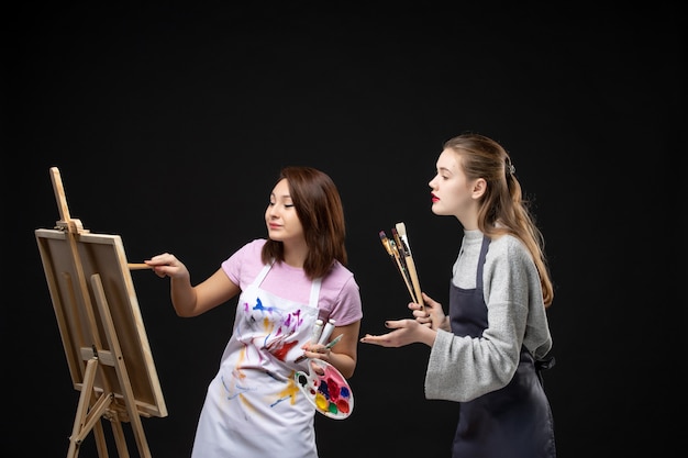 Vista frontal Pintoras dibujando en caballete en la pared negra color dibujar pintura trabajo arte foto artista imagen