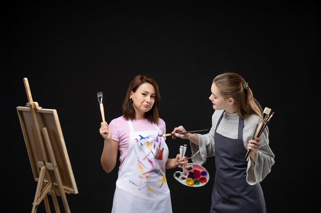 Vista frontal pintoras sosteniendo pinturas y borlas para dibujar en la pared negra dibujar pintura trabajo imagen arte foto color artista