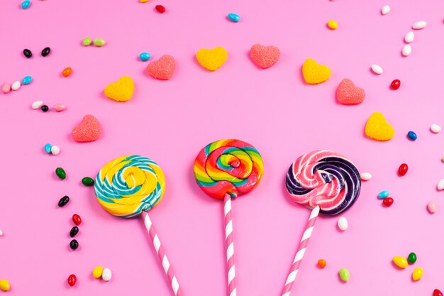 Una vista frontal piruletas de colores sobre palitos de paja dulce de color rosa-blanco junto con mermeladas en forma de corazón y caramelos multicolores en rosa