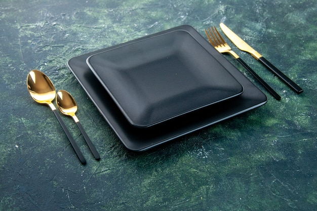 Vista frontal de placas cuadradas negras con cucharas tenedor de oro y cuchillo sobre fondo oscuro
